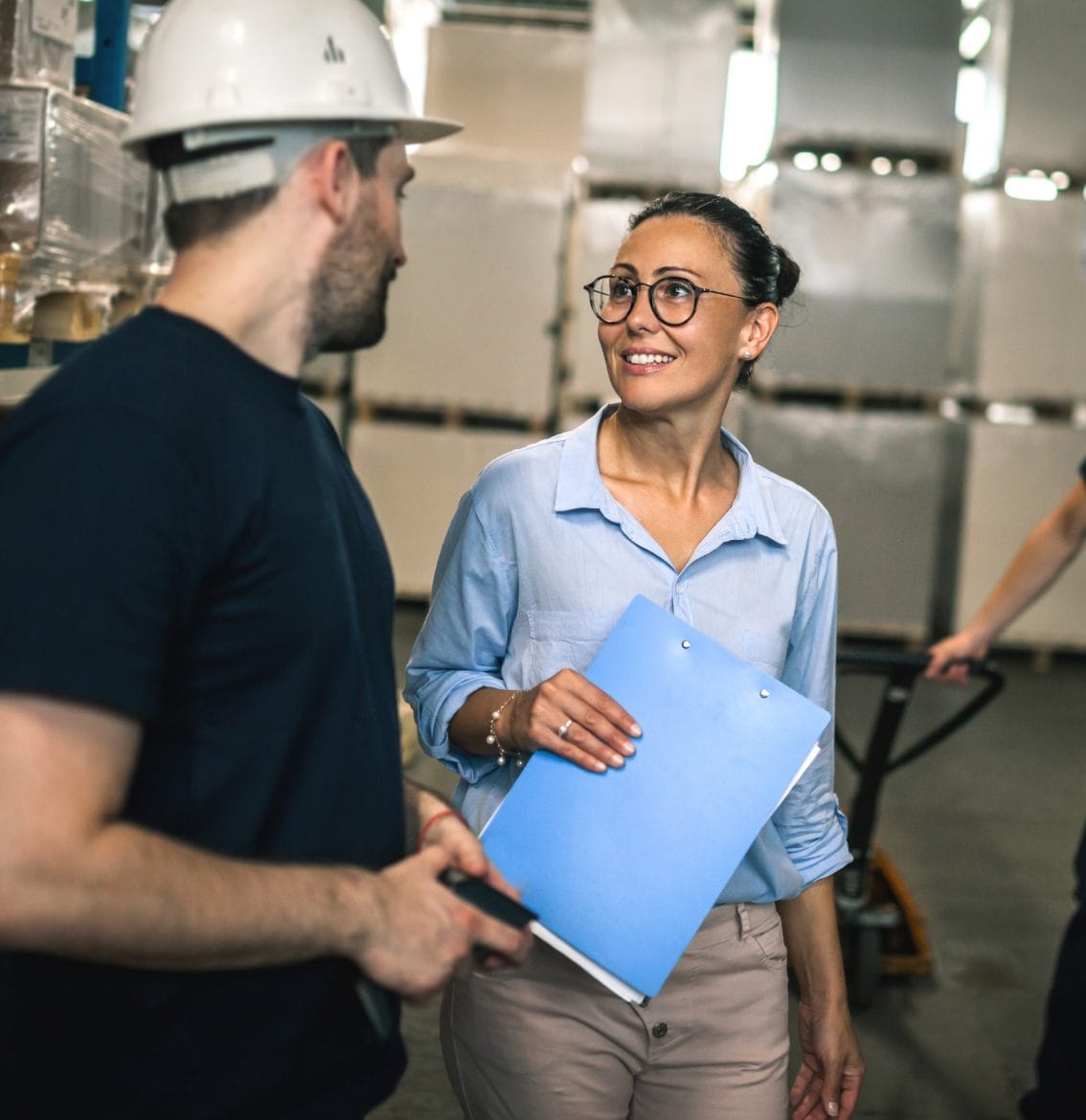 picture of a man in a hardhat talking to a women holding a blue folder talking in a warehouse