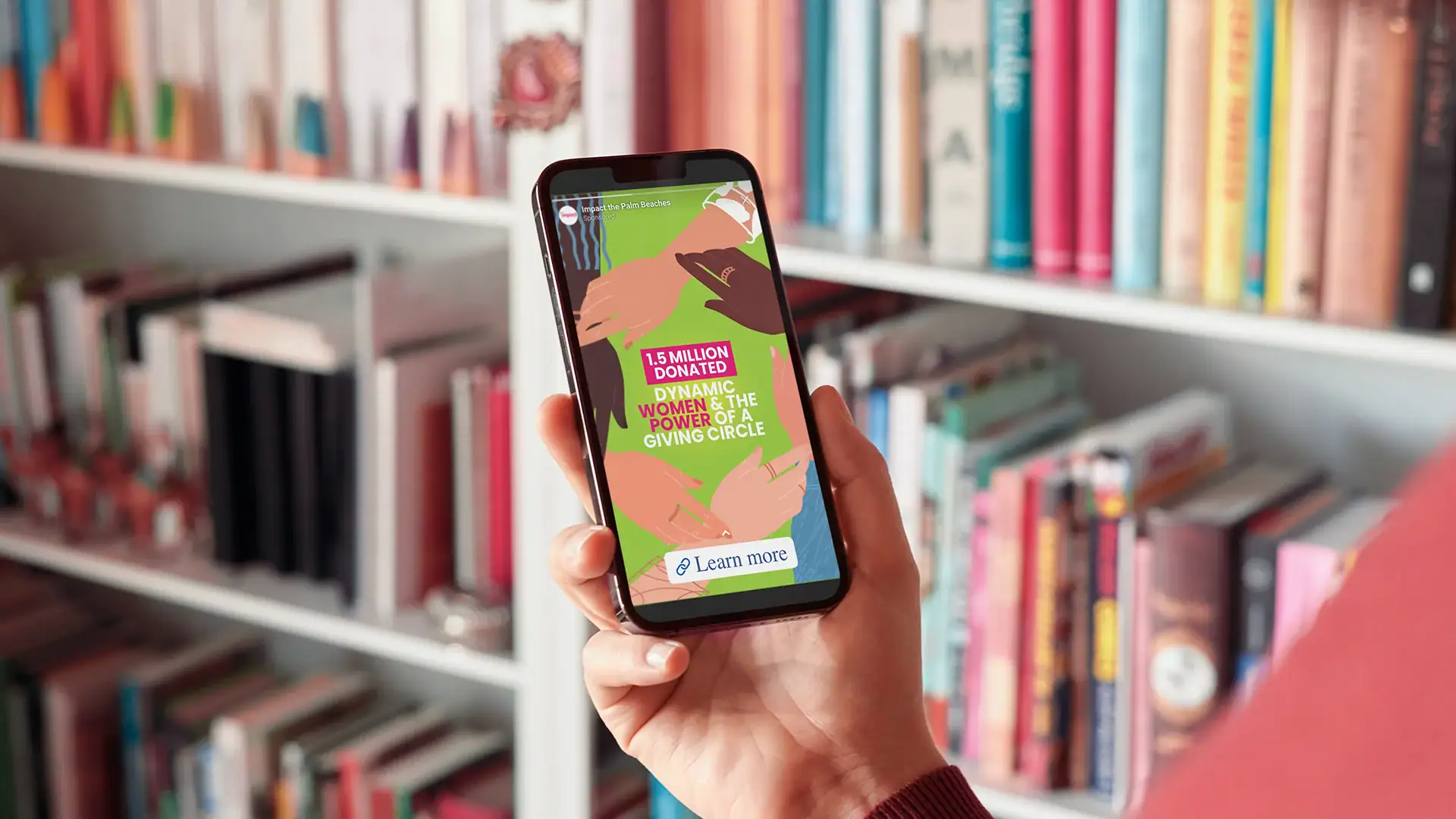Person stands in front of a bookshelf, holding a smartphone displaying Instagram story of Impact The Palm Beaches Philanthropic Giving.